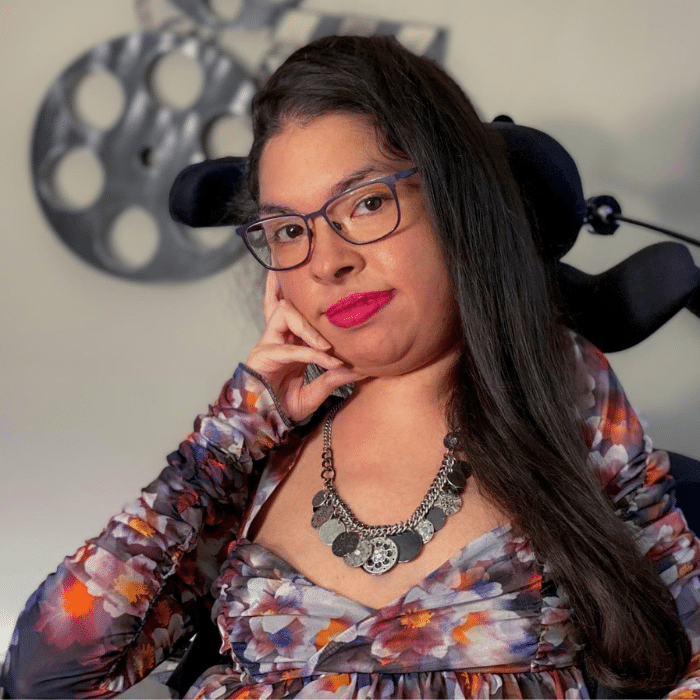 Michelle, a brown gal with long black hair and a hot pink smirk. Her head’s tilted, resting her cheek against her hand and wheelchair headrest. She’s parked in front of a film reel art piece on the wall behind her.