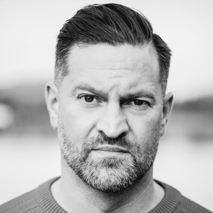 A person with short hair, a beard and a moustache. He is wearing a dark t-shirt. The photo is in black and white.