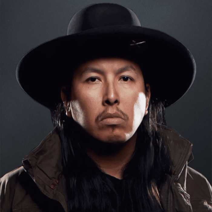 Cowboy Smithx with long, dark hair wearing a wide-brimmed black hat and dark jacket with the collar flipped up.