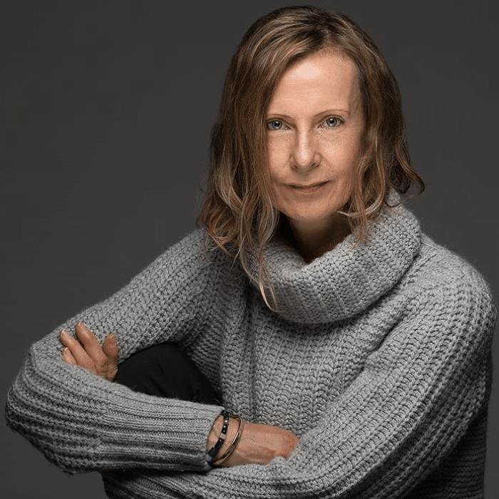 Eva Colmers with brown hair, sitting with arms crossed to the side wearing a grey roll-neck sweater.