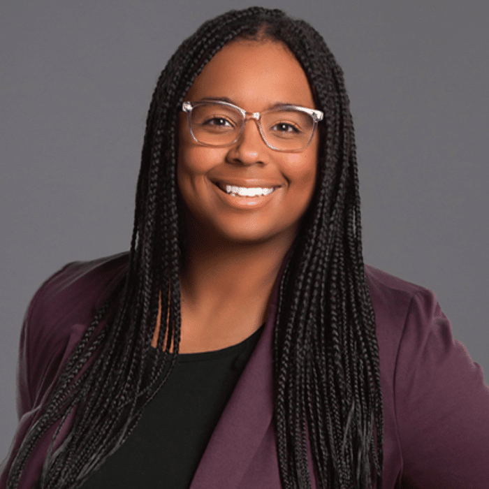 Mariah Braun with long, black box braids, wearing glasses, a plum blazer and a black shirt.