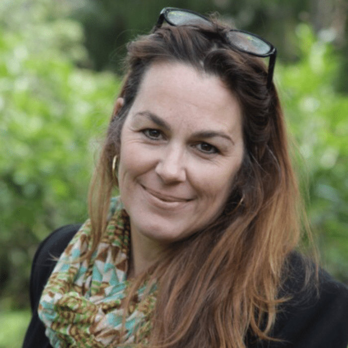 Sarah Robertson with glasses on her head, long brown hair, wearing a turquoise, brown, yellow and white patterned scarf and black shirt.