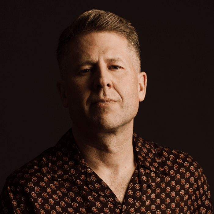 Steve J. Adams partially in shadow with light brown hair, wearing a brown patterned shirt.