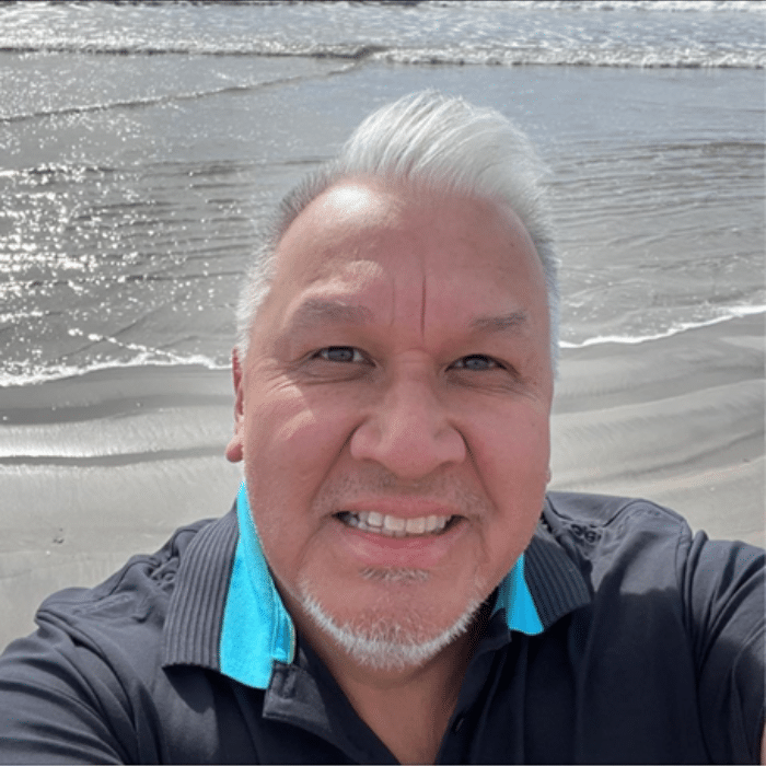 Steve Sxwithul’txw on a beach with short white hair wearing a black shirt with a bright blue collar.