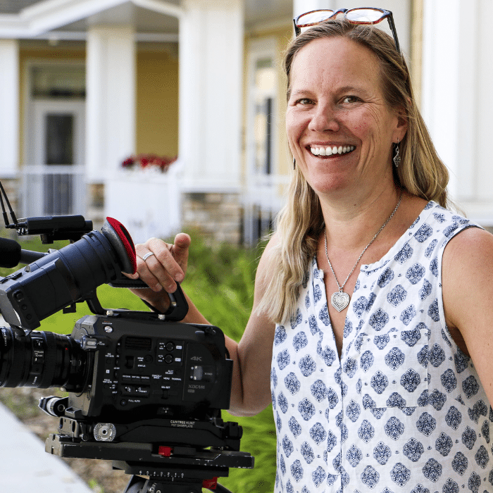 A person with long blonde hair smiles warmly while standing outdoors next to a professional video camera. She is wearing a sleeveless patterned top, with buildings blurred in the background.