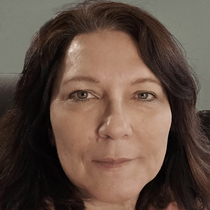 A person with shoulder-length dark hair looks directly at the camera with a neutral expression. She is seated indoors against a plain grey background.
