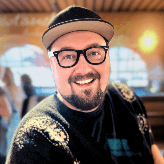 Matt wearing a black baseball cap, black-framed glasses and a black t-shirt with gold and blue embellishments. Matt has a short moustache and beard. He is standing in front of a blurred background.