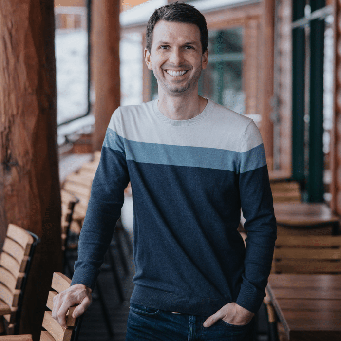 Scott Westby smiling with one hand in his pocket. He is wearing a white, light blue and navy blue blocked long-sleeved shirt and jeans. He is standing in an outdoor wooden seating space.