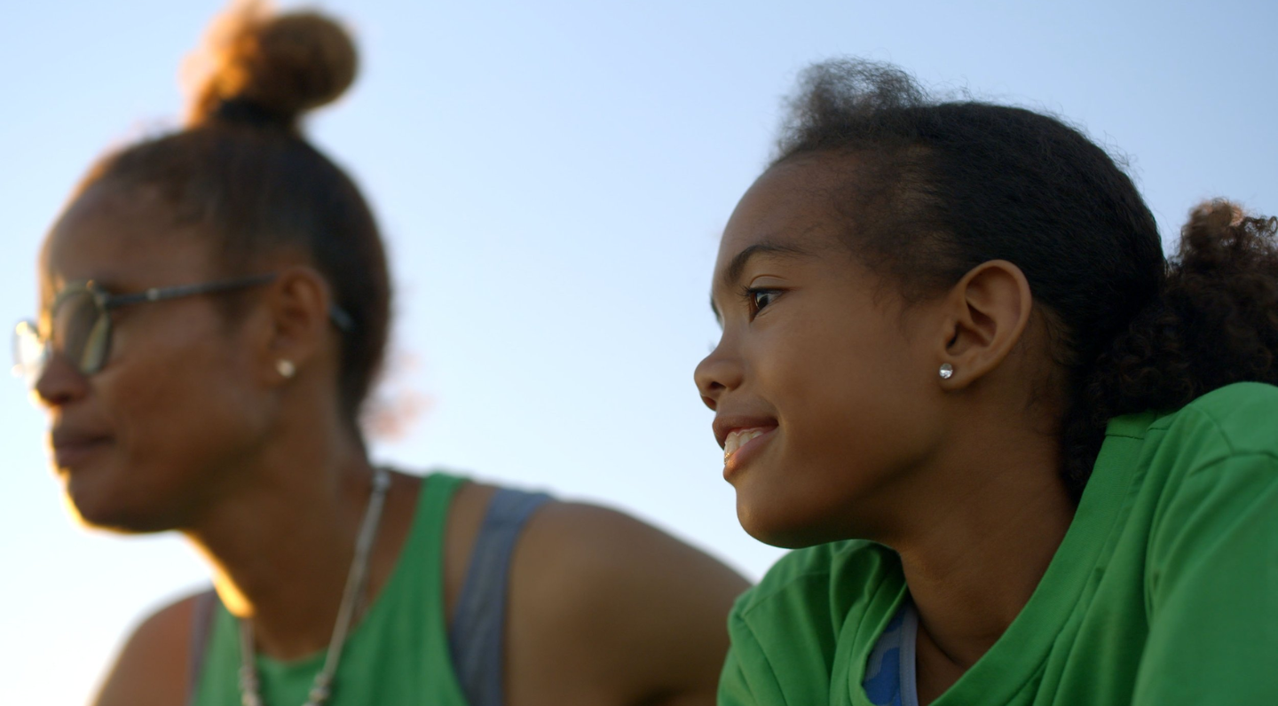 Left side, blurred in background sits a woman with hair piled in a bun on top of her head, wearing glasses, earrings, layered green and grey tank tops and a silver necklace. Right side, foreground, a child with hair in a ponytail, wearing earrings and a green shirt.