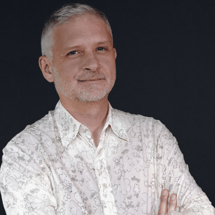 Ian Barr in front of a dark background. He has close cropped white hair and beard / moustache. His arms are crossed. He is wearing a white long-sleeved collared shirt with a black outlined flower pattern.