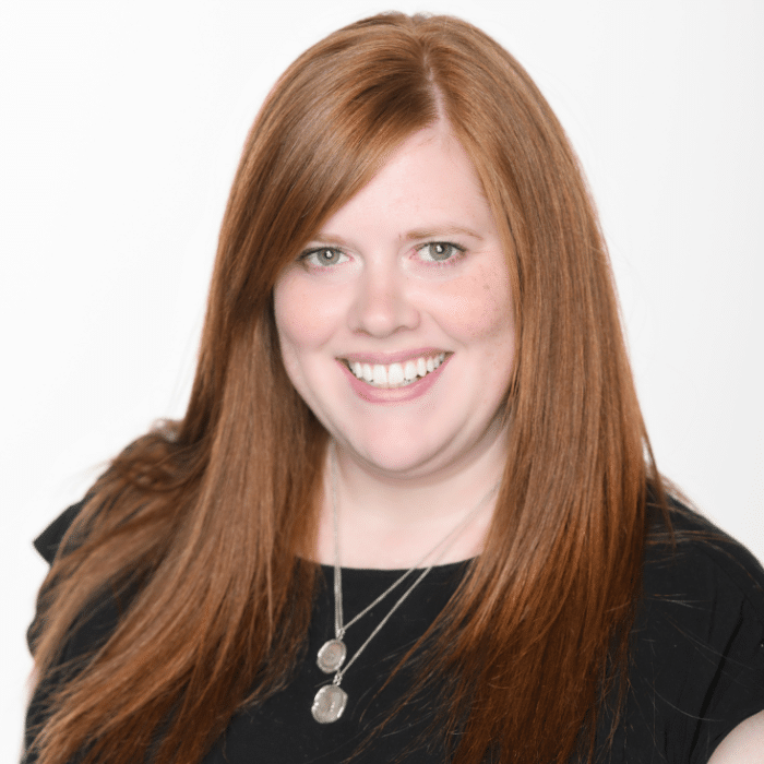 Katrina Beatty is in front of a white background. She has long red hair and is wearing a black short-sleeved shirt with two dangling silver necklace pendants.