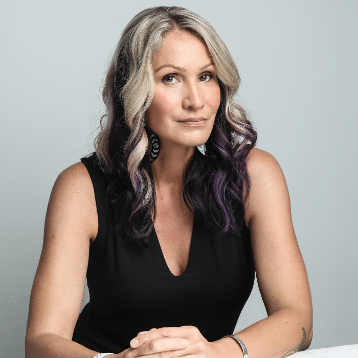 Dr. Jules Koostachin in front of a light grey background leaning with arms resting on a white surface. She has long wavy blonde, black and purple hair. She is wearing black hoop earrings with abalone designs, a black sleeveless shirt and a bracelet on each wrist.