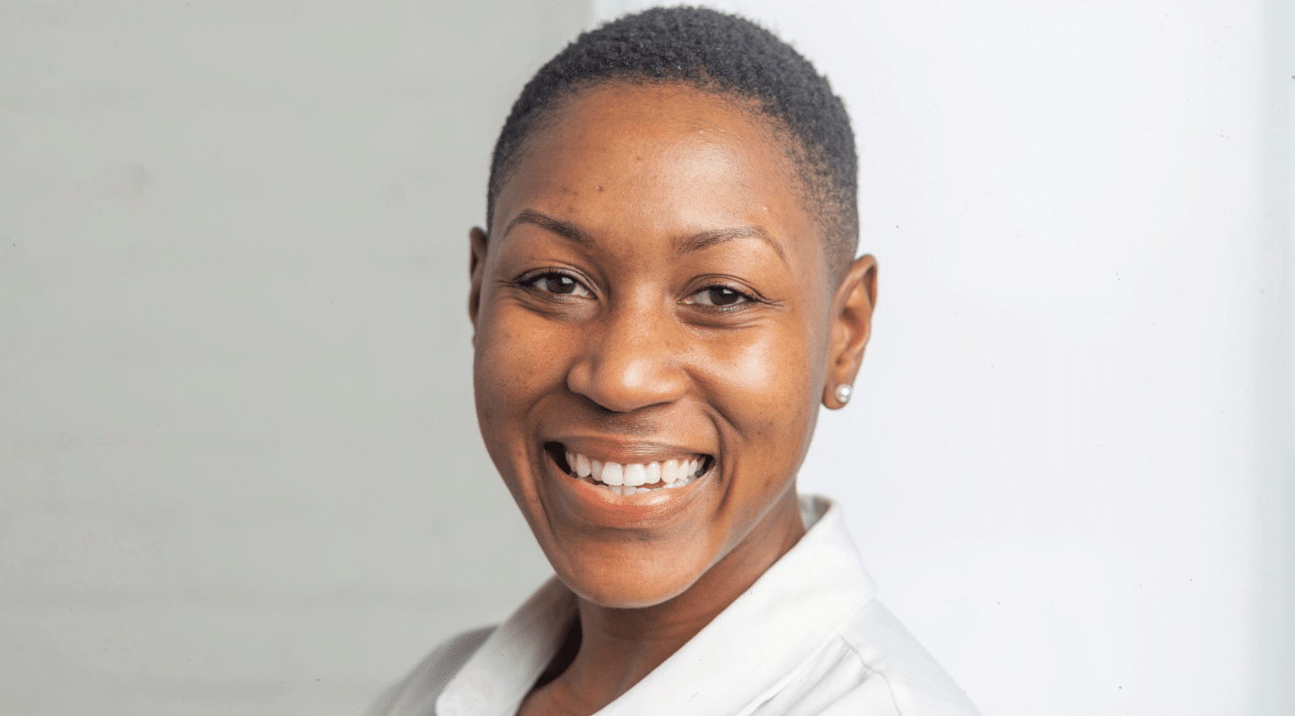 Kema Joseph in front of a grey/white background, smiling. She has close-cropped black hair, is wearing small pearl earrings and a white blouse.