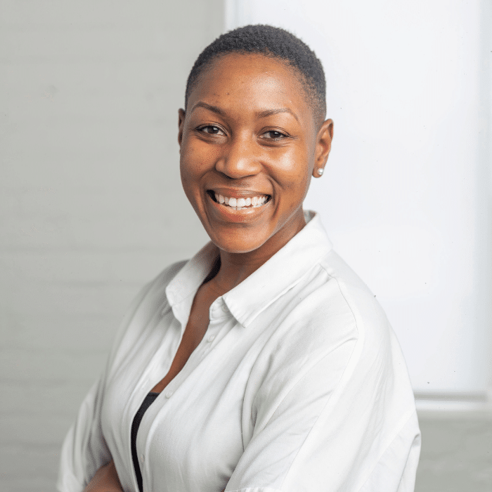Kema Joseph in front of a grey/white background, smiling. She has close-cropped black hair, is wearing small pearl earrings and a white blouse.