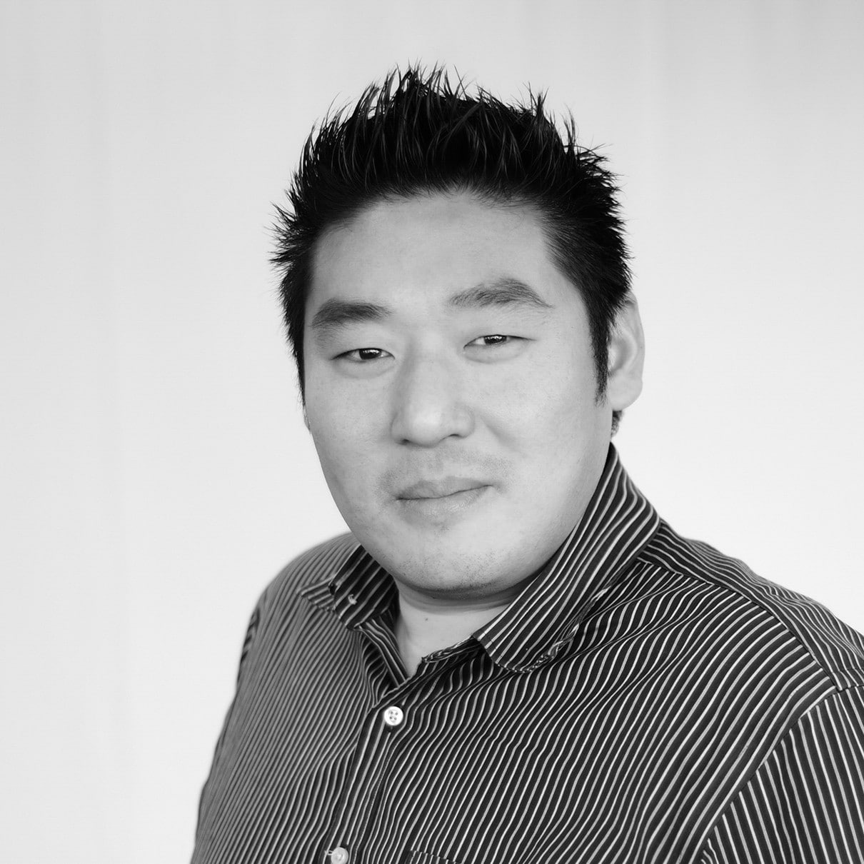 A person with short spiky dark hair. He is wearing a striped, button-up collared shirt. The photo is in black and white.