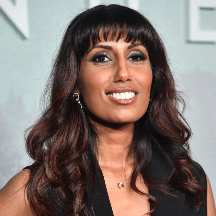 A person with long wavy dark hair smiles. She is wearing a sleeveless black top, silver earrings and a silver necklace, photographed against a pale blue-grey backdrop with blurred letters.