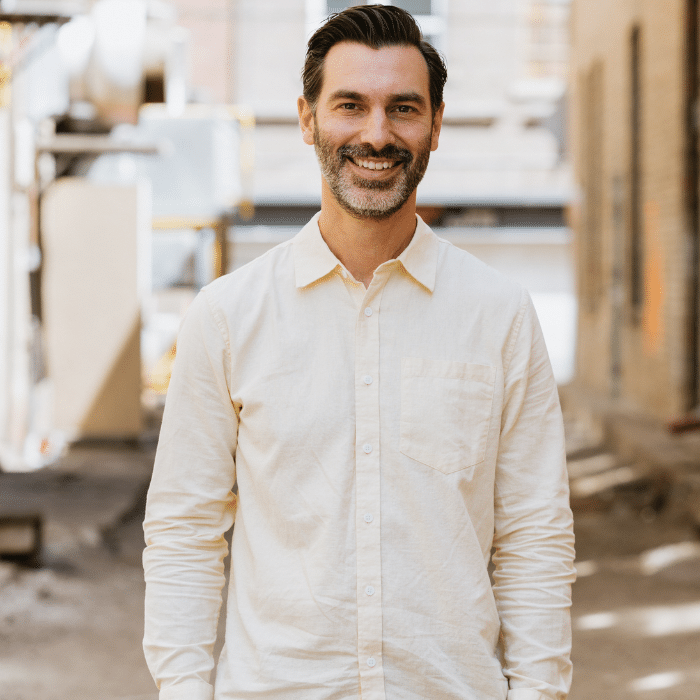 Kyle Burgess standing in front of a blurred background. He has dark hair, salt and pepper facial hair. He is wearing a long-sleeve off-white dress shirt.