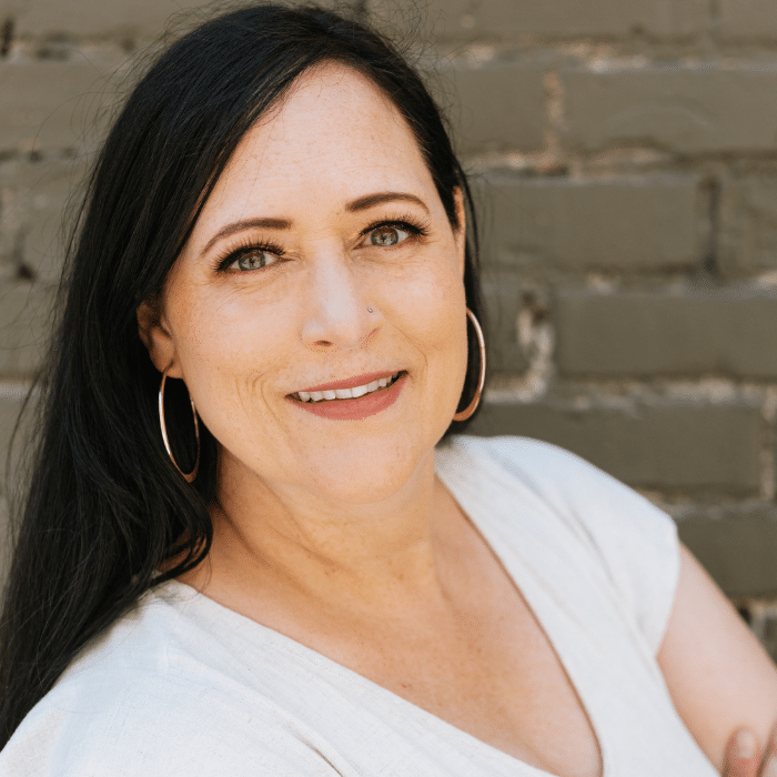 Allia in front of a grey brick wall. She has long dark hair parted on the side. She is wearing hoop earrings and a white v-neck t-shirt with her arms.
