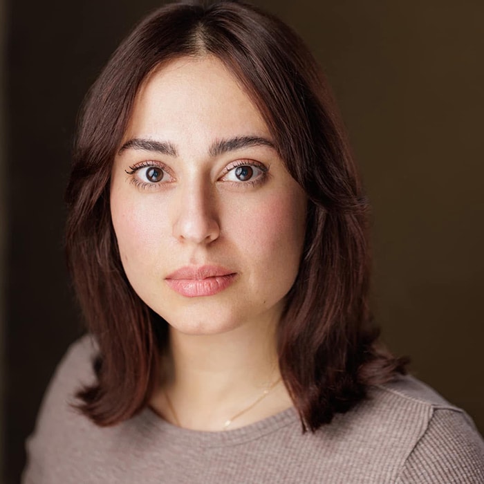 A professional headshot of a woman with shoulder-length brown hair, soft waves framing her face and natural makeup. She has fair skin, expressive brown eyes and is wearing a neutral-toned top. The background is a muted, warm brown colour, providing a simple and elegant setting. Her expression is calm and confident, with a direct gaze toward the camera.