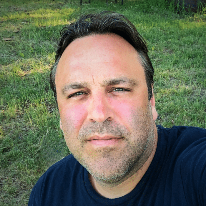 Douglas has short dark hair, stubbly facial hair and is wearing a navy blue crew neck t-shirt. There is a grassy field in the background.