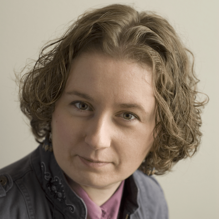 Kelly-Anne has short wavy brown hair. She is wearing a mauve shirt underneath a grey jacket with an embroidered design on the collar.
