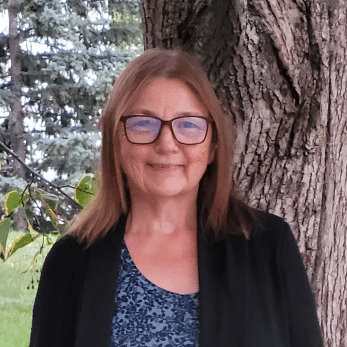 Judith has shoulder-length light brown hair. She is wearing glasses, a blue patterned shirt and a black cardigan. Judith is standing in front of a tree.