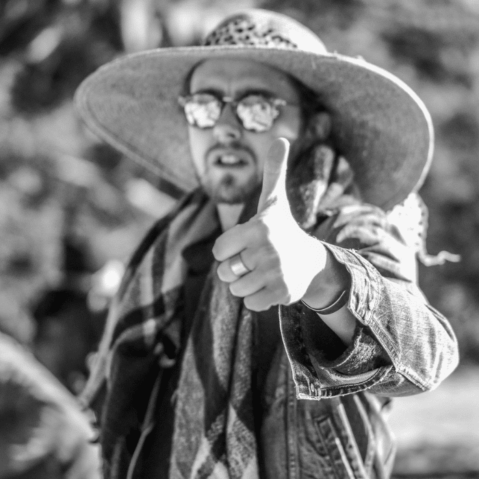 Black and white image of Style Dayne Stenberg giving a thumbs up. He is wearing a wide-brimmed hat, mirrored sunglasses and a jean jacket with a plaid scarf around his neck.