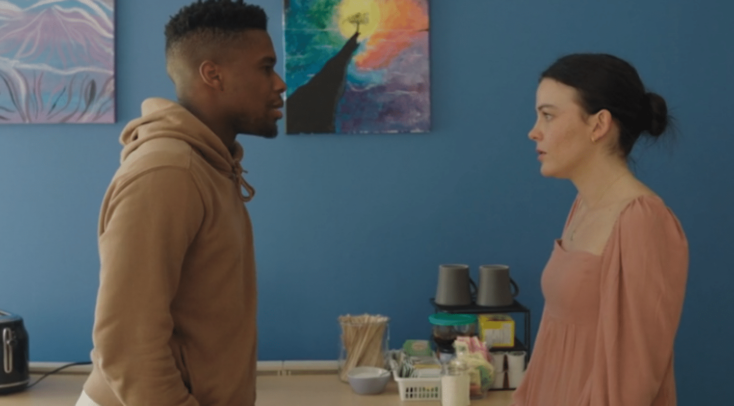 A Black man and a white woman stand facing each other in a breakroom, appearing to be in the middle of an intense conversation. The man, wearing a tan hoodie, looks directly at the woman, while she, dressed in a muted pink dress, looks back at him with a calm but focused expression. Behind them, a counter holds mugs, tea supplies, and a kettle, with colorful abstract artwork hanging on a bright blue wall.