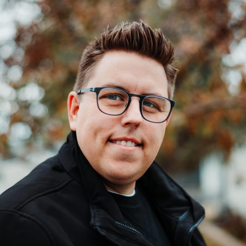 A person with short, spiked brown hair and glasses smiles slightly while looking off to the side. They are wearing a black jacket, and the background is softly blurred with autumn-coloured leaves.