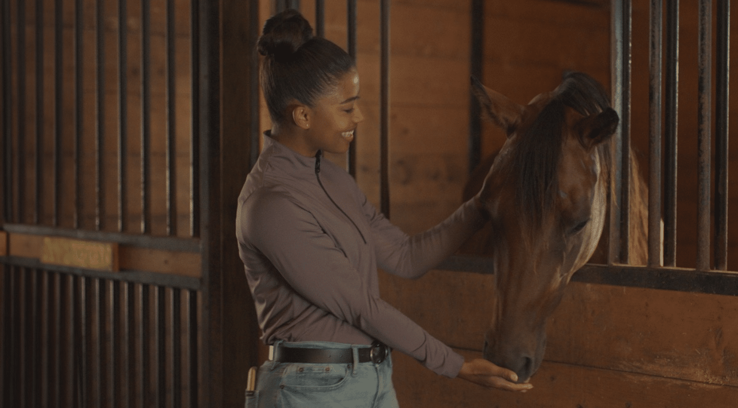 A woman smiles while feeding a brown horse inside a stable. She is wearing a mauve zip-up top and jeans, with her hair in a high bun. The background shows wooden stall walls and vertical black bars.
