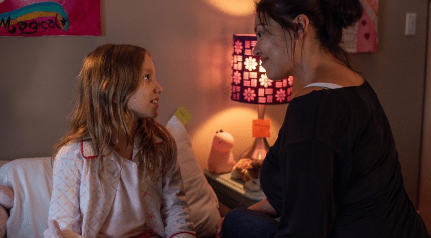 A young girl in pajamas sits on a bed talking with an adult who is sitting beside her. They are warmly lit by a pink lamp on a nightstand, which is surrounded by plush toys and drawings.