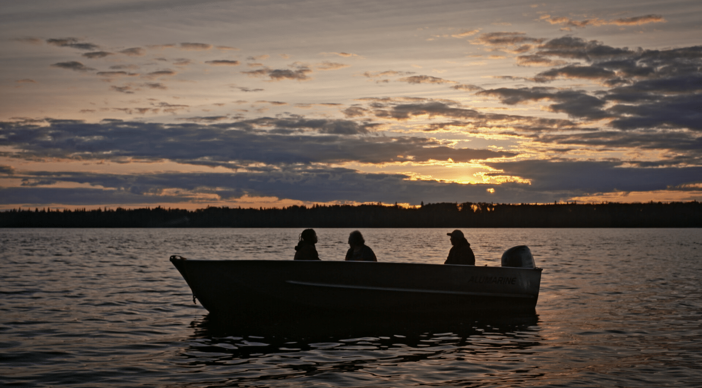 Three people sit in a small motorboat on a lake at sunset. The boat is silhouetted against a sky filled with clouds and warm orange and purple tones. A dark treeline is visible on the horizon.