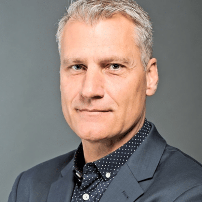 A person with short gray hair wearing a dark suit jacket and navy shirt with white polka dots, posing against a plain gray background.