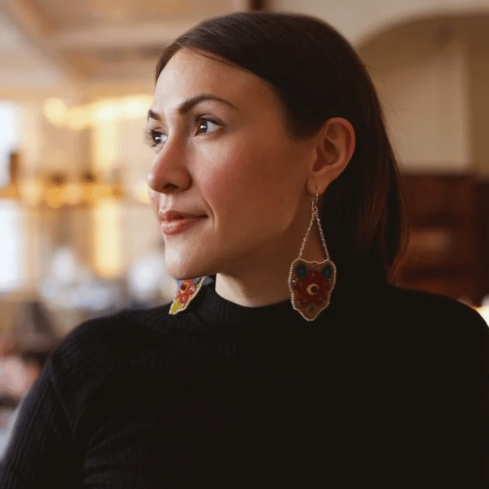 An Indigenous person with long dark hair wearing a black sweater and colourful beaded earrings, looking to the side in an indoor setting with a blurred background.