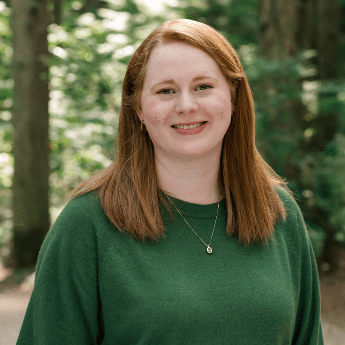 A person with shoulder-length straight red hair smiles softly at the camera. She is wearing a green sweater and a thin gold necklace, standing in a forested outdoor setting.