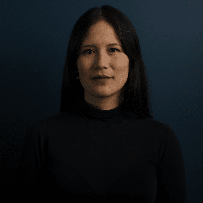 A person with long dark hair stands against a deep blue background. She is wearing a black turtleneck.
