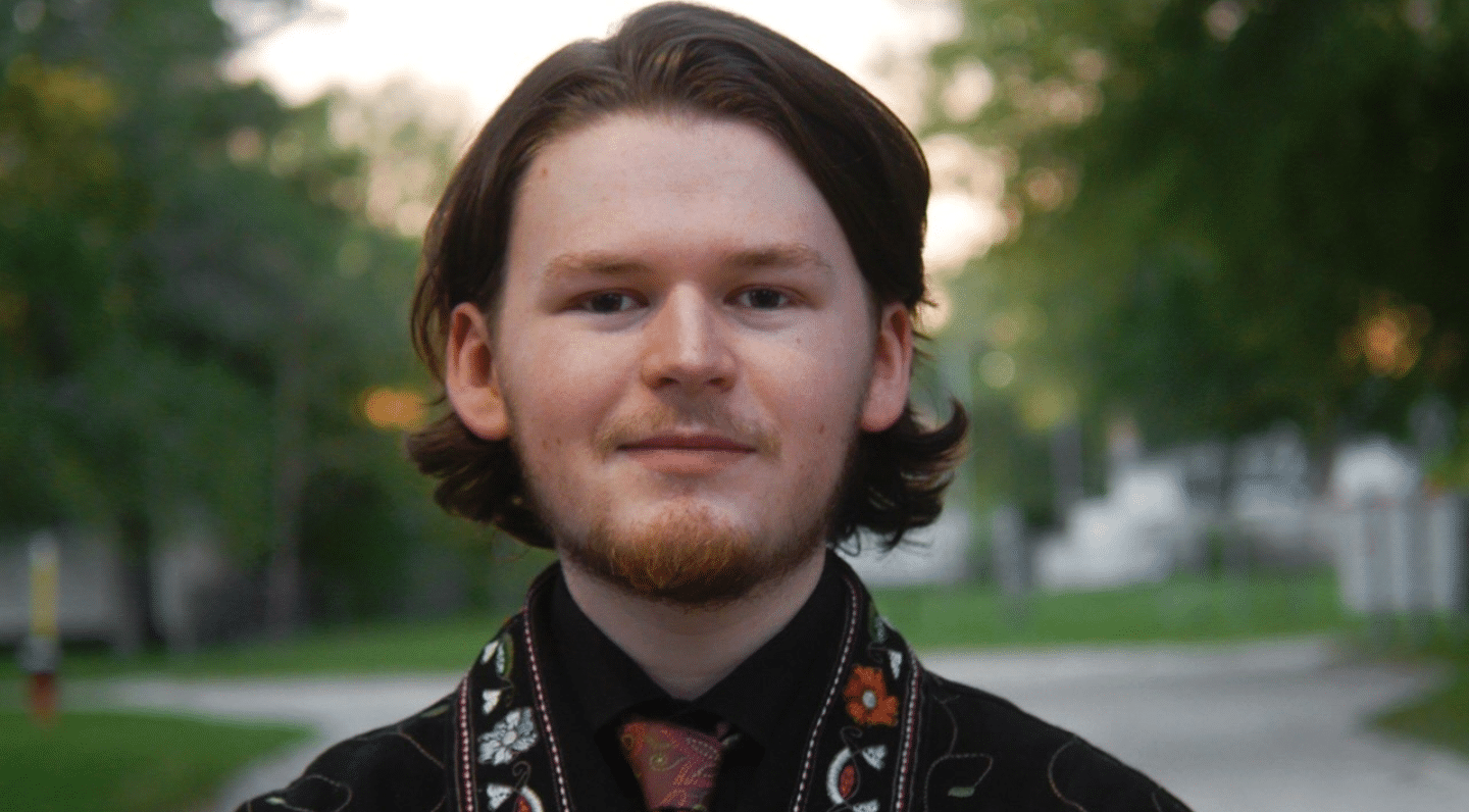A person with chin-length brown hair and a short beard standing outdoors on a pathway with a neutral facial expression. He is wearing a black shirt with an embroidered floral-patterned vest and a patterned tie. Trees and greenery are blurred in the background.