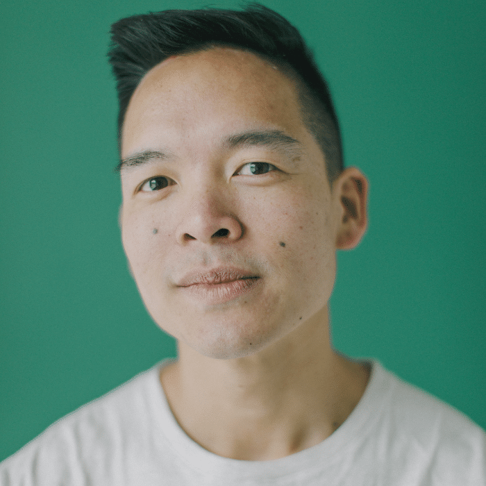 A person with short dark hair and a neutral expression poses against a solid green background, wearing a light-colored shirt