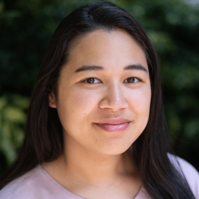 A person with long dark hair smiles at the camera. She is wearing a light pink top, and the background is filled with green foliage.