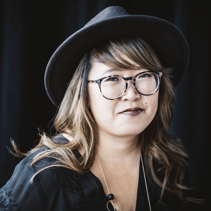 A person with wavy light-brown hair wears a wide-brimmed dark hat and tortoise shell-framed glasses. She is wearing dark lipstick, a long necklace and a black top, posed against a dark backdrop.