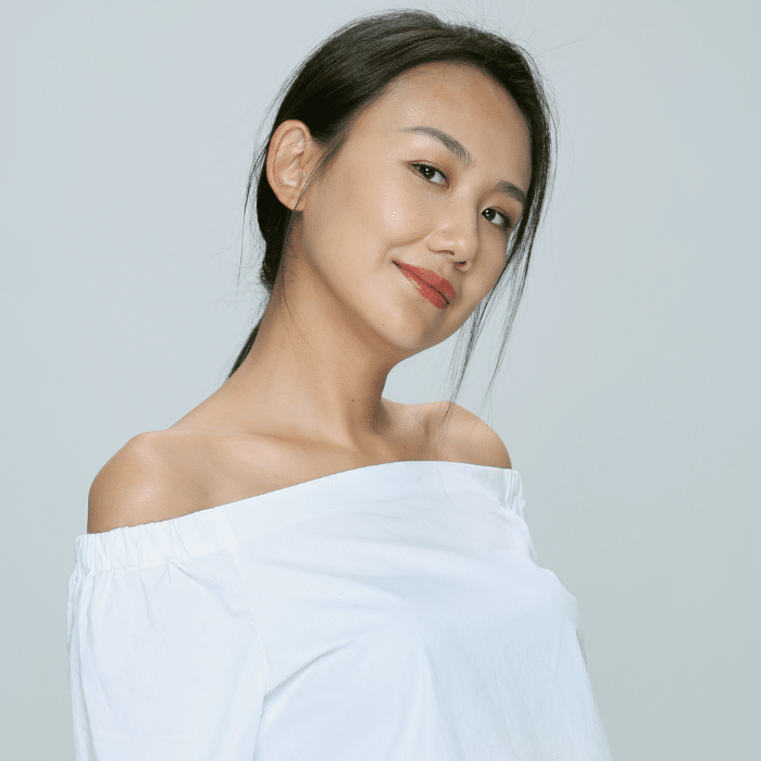 A person with long dark hair loosely pulled back smiles. She is wearing an off-the-shoulder white top. The background is a smooth pale grey.