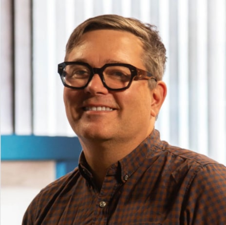 Colour portrait of a man with short brown hair and black-framed glasses, smiling slightly at the camera. He is wearing a brown patterned button-up shirt. The background features vertical light panels and a teal accent, suggesting a studio or office setting.