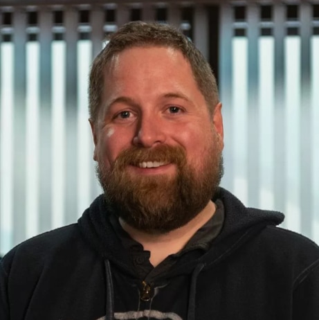 Colour headshot of a man with short hair and a full beard, smiling warmly at the camera. He is wearing a dark hoodie over a T-shirt. The background includes vertical light panels, softly out of focus, indicating an indoor studio or workspace environment.