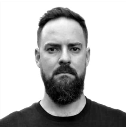 Black-and-white headshot of a man with short dark hair and a full beard, facing the camera with a neutral expression. He is wearing a dark crew-neck shirt. The background is plain and light coloured, with no visible details.